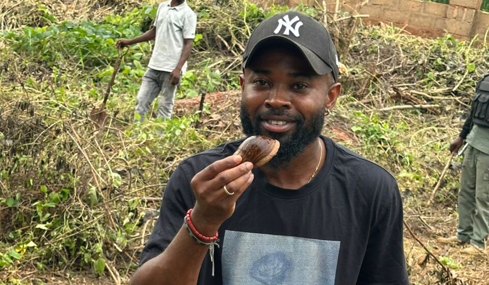 Ein Mann, der ein schwarzes T-Shirt und eine schwarze Mütze trägt, hält einen braunen Gegenstand, möglicherweise eine Nuss, in die Höhe, während er in einem Außenbereich mit überwucherter Vegetation und anderen Menschen im Hintergrund steht und auf eine Initiative von Spende Nigeria hinweist.